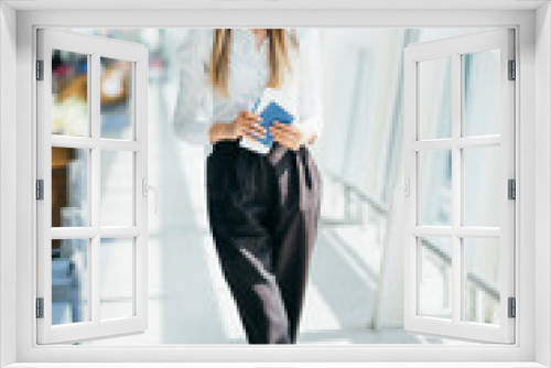 Young girl, business woman standing by the window in a modern airport holding tickets and a phone, looking at the camera. Portrait of a beautiful girl in a mask. Business trip by plane