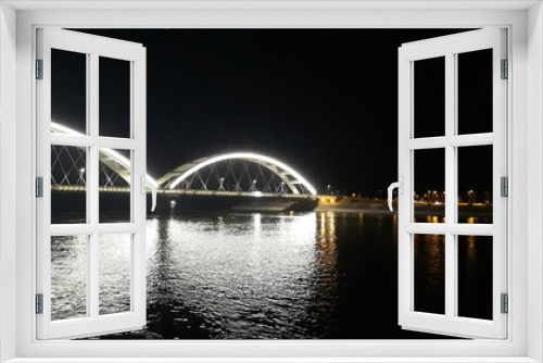 Fototapeta Naklejka Na Ścianę Okno 3D - Die Brücke über den Fluss wirft Weiß in die Nacht. Die Lichter der nächtlichen Stadt spiegeln sich im Fluss. Silhouetten von Siedlungen am See. Verschwommene Fotos in der Dunkelheit der Nacht.