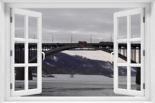 Fototapeta Naklejka Na Ścianę Okno 3D - Landscape with a span of a large arched bridge and a frozen river. A tram and a bus have met on the bridge and cannot separate. A tram with an empty passenger compartment is moving over a beautiful br