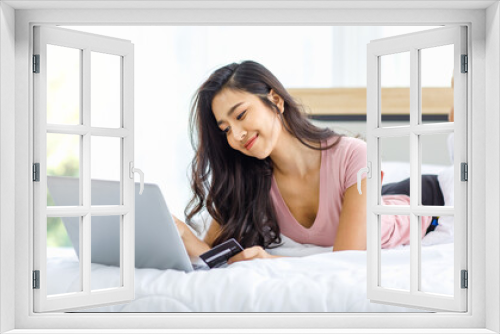 Asian young happy cheerful beautiful female model in casual pajama outfit lying down smiling hold credit card on bed in bedroom browsing surfing internet shopping online via laptop notebook computer