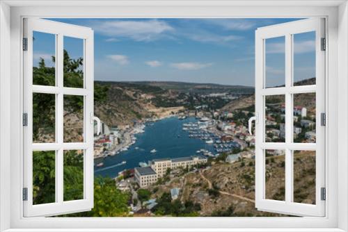 Fototapeta Naklejka Na Ścianę Okno 3D - View from the mountain to the city with a pier and a bay. City in the mountains. Around the green foliage, trees, blue sky. Yachts, boats, buildings, houses are visible in the city. Urban landscape.