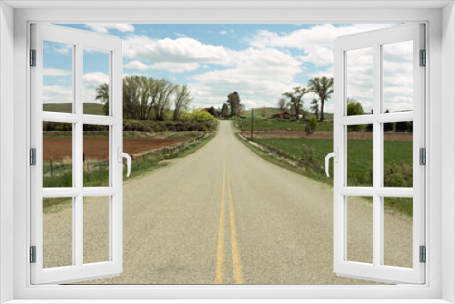 Fototapeta Naklejka Na Ścianę Okno 3D -  Looking down middle of the road in Shell, Wyoming, you see pasture land, trees and farm houses with the horizon and blue sky and clouds beyond.