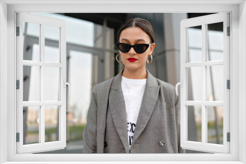 Incredible stylish woman with dark hair and red lips wearing golden earrings and grey jacket posing at camera outdoor against glass building