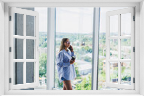 Modern woman freelancer talking on the phone while standing at a large window and drinking coffee. The concept of a modern successful woman. Young girl in an open office