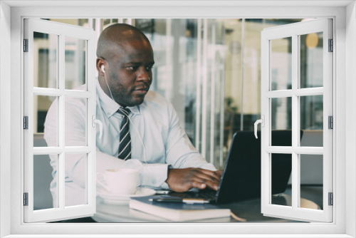 African American businessman in formal wear working and surfing social media on his laptop while listening to music with earbuds in a glassy cafe