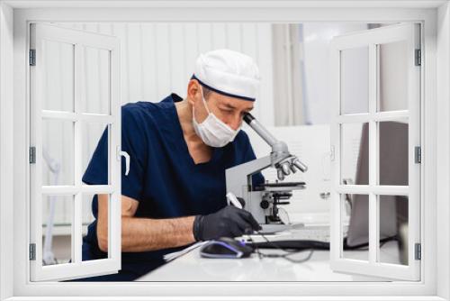 A male doctor in blue lab suit, white hat, mask and black gloves studies a stained histology slide with a microscope in the lab