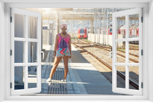 A girl in summer clothes with a backpack waiting for an electric commuter train arriving in the distance.