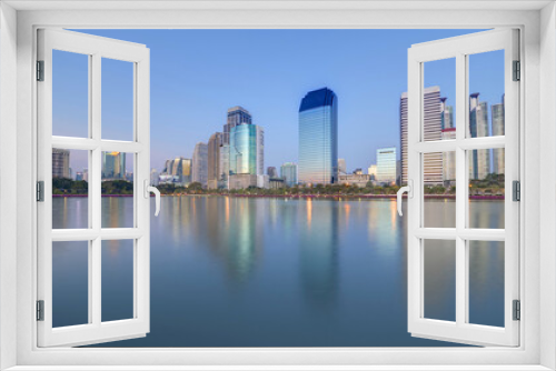 Fototapeta Naklejka Na Ścianę Okno 3D - Night skyline of modern lakeside skyscrapers with glass curtain walls and dazzling city lights reflected in the smooth lake water in beautiful Benjakiti Park at blue dusk, in Bangkok, Thailand, Asia