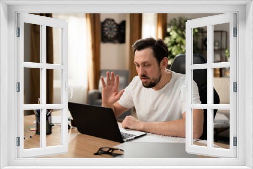 A young man holds conference on his laptop. He greets his colleagues and shares the results of work. Stationery and documents are on the table.