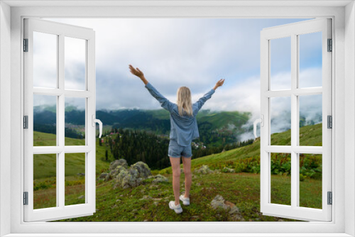 Fototapeta Naklejka Na Ścianę Okno 3D - Young slender blonde woman in shorts and a shirt stands with her hands raised up on a mountain above the clouds. Bakhmaro, Georgia