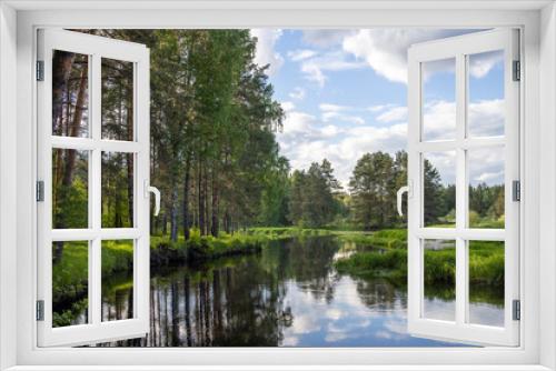 Fototapeta Naklejka Na Ścianę Okno 3D - Bright sunny landscape with pine trees near the river. The sun's rays illuminate the young greenery and trees. The sky and clouds are reflected in the river.
