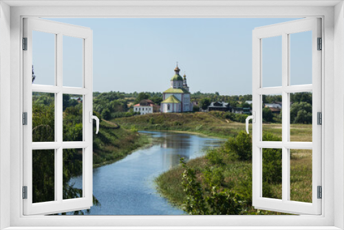 Fototapeta Naklejka Na Ścianę Okno 3D - The white stone Church of Elijah the Prophet on the bank of the Kamenka river in Suzdal Russia on a summer day and old houses among lush green foliage