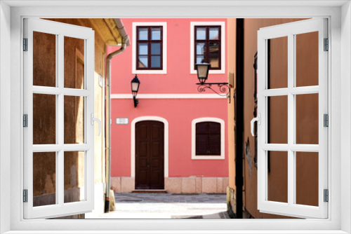 Fototapeta Naklejka Na Ścianę Okno 3D - narrow alley way in European town with yellow and pink stucco walls and facades. old style wood windows. travel and tourism concept. classic european residential architecture. sconce street lighting.