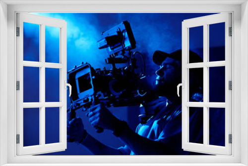 Fototapeta Naklejka Na Ścianę Okno 3D - Young videographer in casualwear shooting commercial video in dark room or studio full of smoke lit by dark blue light