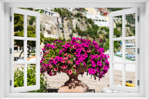 Fototapeta Naklejka Na Ścianę Okno 3D - A pot of purple flowers sitting on a ledge in direct sunlight with cliffside Mediterranean architecture in the background in Positano, Italy, Europe.