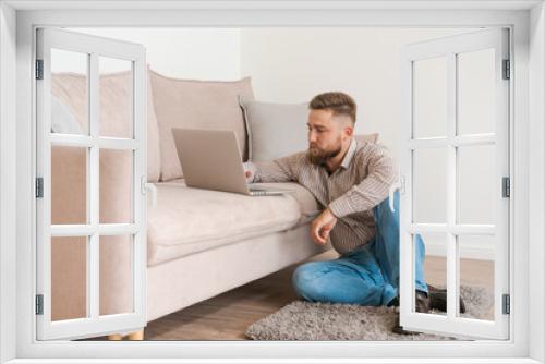 Young attractive, smiling bearded guy browsing his laptop while sitting at home on a cozy beige sofa at home, in casual clothes on a day off