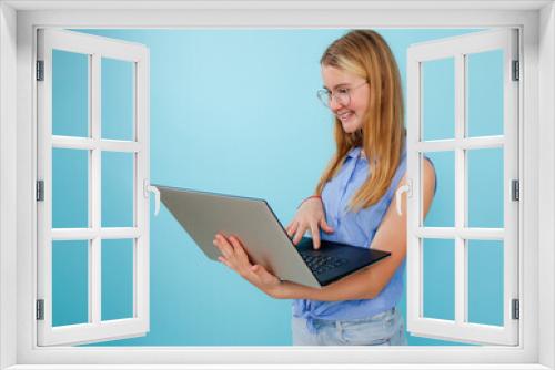 Smiling, confident, successful blonde woman in glasses holding and using laptop on blue isolated background. Freelancer