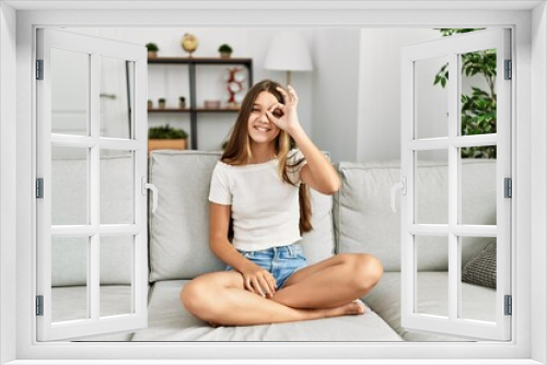 Young brunette teenager sitting on the sofa at home doing ok gesture with hand smiling, eye looking through fingers with happy face.