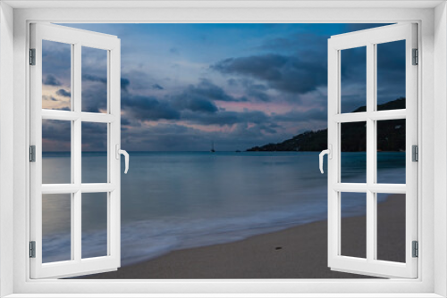 Fototapeta Naklejka Na Ścianę Okno 3D - A quiet evening on a tropical beach. The waves of the turquoise ocean are foaming on the sand. Clouds in the sky, highlighted in pink. A hill in the distance. Reflection on the water. Long exposure.