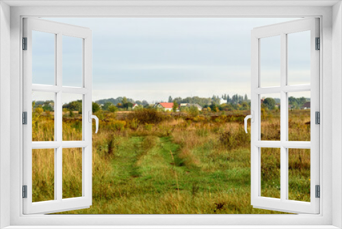 Fototapeta Naklejka Na Ścianę Okno 3D - The picture shows an autumn natural landscape, a wide field with yellowing grass, roofs of houses on the horizon and a blue sky.