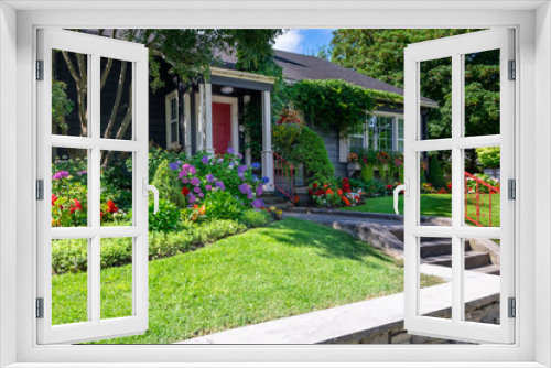 Fototapeta Naklejka Na Ścianę Okno 3D - The front yard of a dark blue house with a vibrant red door. The building has a glass window with white trim. The colorful garden has green grass, red, pink, and yellow flowers, shrubs, and trees. 