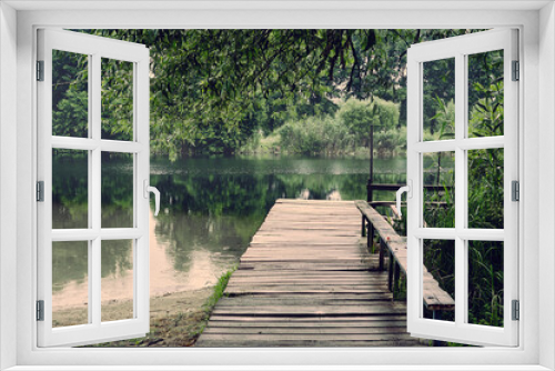 Fototapeta Naklejka Na Ścianę Okno 3D - It's cloudy, it's raining a little, an old wooden pier approaches a small river, grass and trees are all around, a beautiful rural landscape
