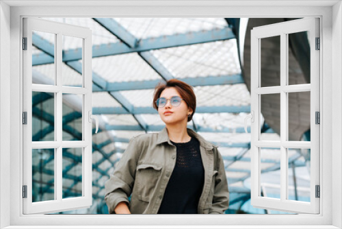 Fashionable young woman in fashion round glasses posing on background of modern architecture looking away. Beautiful asian woman with short haircut standing outdoors, low angle view portrait