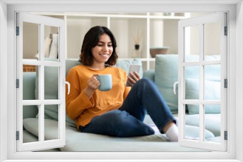 Weekend Leisure. Smiling Arab Woman Relaxing On Couch With Smartphone And Coffee