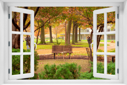 Fototapeta Naklejka Na Ścianę Okno 3D - Outdoor seating on a college campus. Trees overhang the space with green, orange and red fall leaves. Students in coats walk by. More trees and a building in the background.