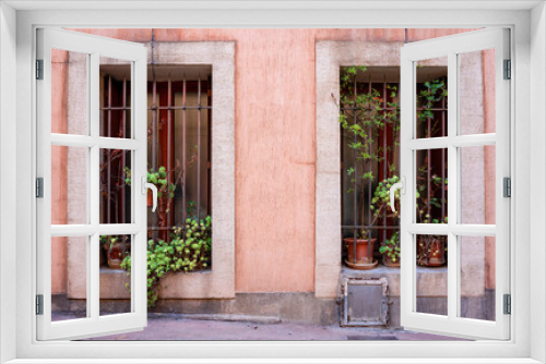 Fototapeta Naklejka Na Ścianę Okno 3D - Frontal view of a pink traditional house facade in a small French village. two large windows with bars and flower pots. Green plants hang in the window