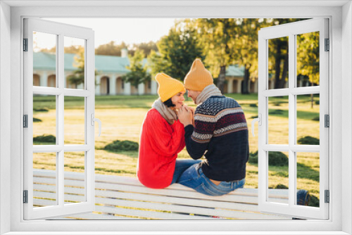 Fototapeta Naklejka Na Ścianę Okno 3D - Romantic couple sit on bench, enjoy sunny day, keep hands together, look with great love at each other, have good relationships. Female has date with boyfriend in park, admire beautiful nature