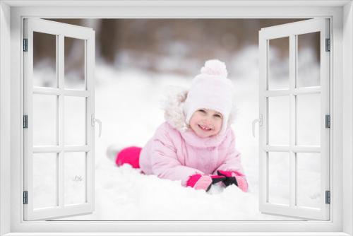 Smiling beautiful little girl lying down on white snow at city park. Cute 3 years old toddler enjoying cold winter day. Front view. Closeup. Posing and looking at camera.