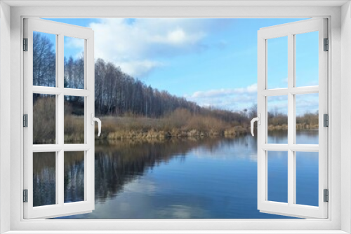 Fototapeta Naklejka Na Ścianę Okno 3D - The sky over the lake in November is blue with feathery clouds. Dry grass stands on the shore, bushes and trees grow. The wind has created some ripples on the water. The weather is sunny and cold