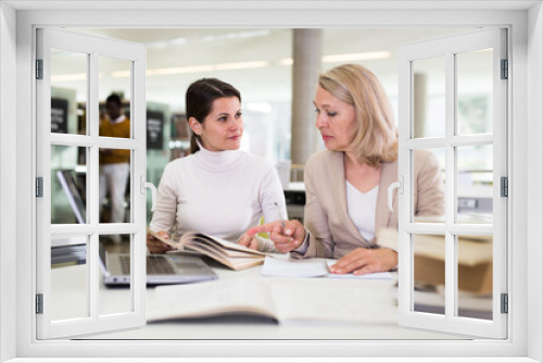 Friendly female tutor helping to diligent positive girl preparing for exam in library. High quality photo