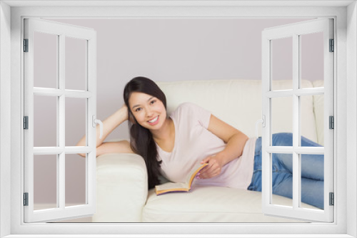 Happy asian girl lying on the sofa reading book smiling at camer