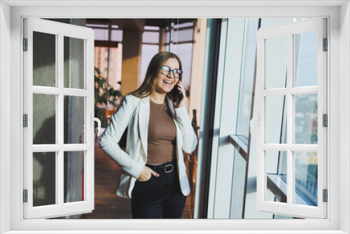 A fair-haired European woman in a white jacket and glasses is talking on the phone while standing in a spacious office with large windows. Successful young woman freelancer