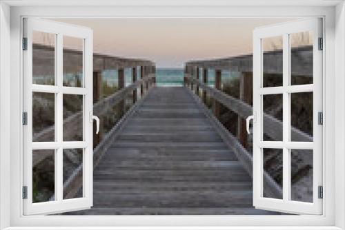 Fototapeta Naklejka Na Ścianę Okno 3D - Vertical shot view of a wooden pathway with railings in between grassy sand dunes in Destin, Florida. Pathway heading to the beack with blue ocean under the horizon skyline.
