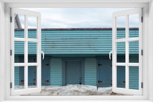 Fototapeta Naklejka Na Ścianę Okno 3D - A blue color exterior wooden wall of a vintage warehouse or garage with multiple double doors and a single wooden door in the middle. The wooden doors are a darker shade of blue. It's winter with snow