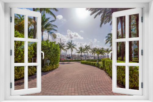 Fototapeta Naklejka Na Ścianę Okno 3D - Stone bricks pavement with bushes and palm trees on the side at Miami River Walk- Miami, Florida. Pathway with gate on the left and a view of boats at the background under the sun.