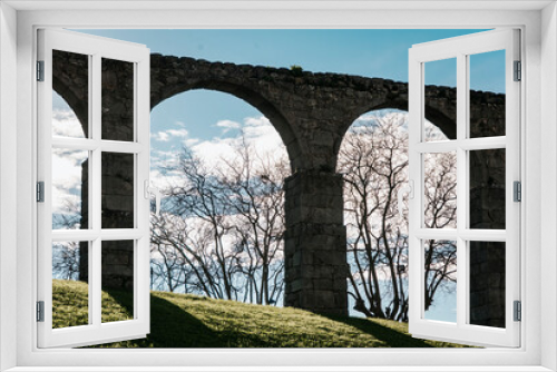 Fototapeta Naklejka Na Ścianę Okno 3D - Arches of the Santa Clara Aqueduct that surrounds the church in Vila do Conde, Portugal seen from below, backlit with dry trees, lawn and clouds in the sky