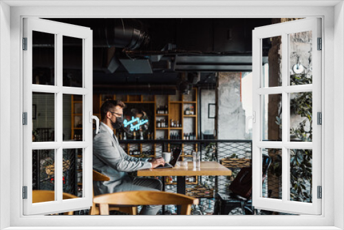 A businessman with a protective mask on his face is sitting in a cafe and talking on the phone while using laptop. Coronavirus, Covid-19 concept.