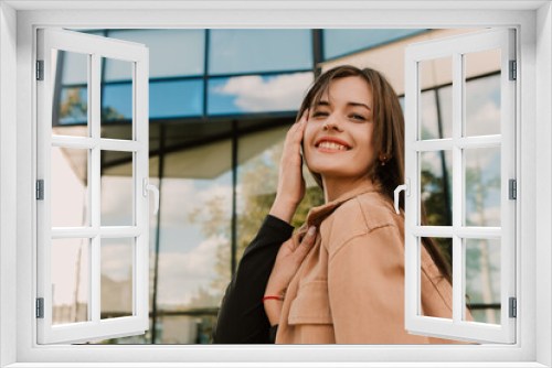 Caucasian young pretty stylish woman standing near business office center street dressed brown trench coat smiling happy poses outside in city, spring autumn season. Cute brunette hair lady
