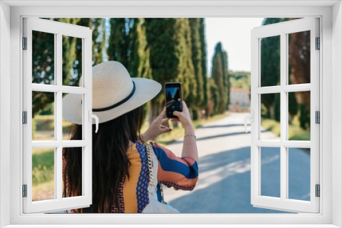 Fototapeta Naklejka Na Ścianę Okno 3D - Back view of woman in straw hat taking photos of  idyllic road in Tuscany with mobile phone