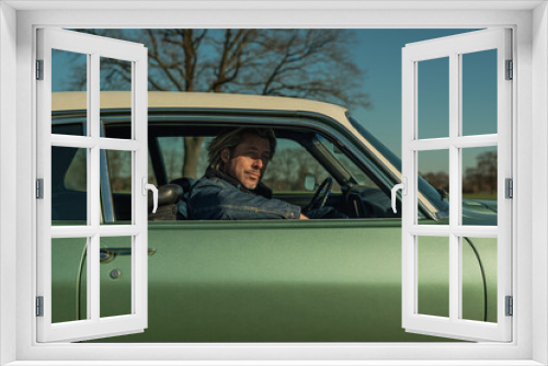 Man with blond hair in jeans jacket sits on passenger side of a vintage american muscle car looking out the window.