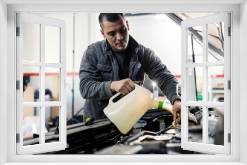 An auto mechanic is pouring oil into the car engine under the hood while standing at garage.