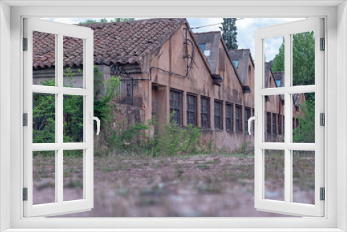 Fototapeta Naklejka Na Ścianę Okno 3D - Abandoned factory with sawtooth-shaped roofs and barred windows. Dilapidated and overgrown, highlighting the passage of time. Eerie beauty captures the mystery of industrial decay.