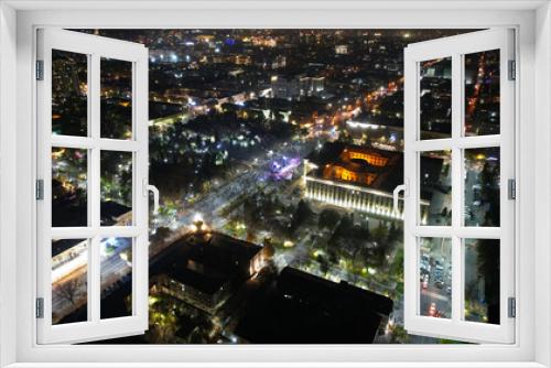 Fototapeta Naklejka Na Ścianę Okno 3D - A big concert in the night city. Drone view of a crowd of people, rays and lights of light. A large building with lighting. There are roads with cars, pedestrian streets with people and a park around