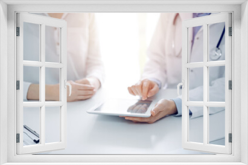 Doctor and patient sitting near each other at the desk in clinic. The focus is on female physician's hands pointing into tablet computer touchpad, close up. Medicine concept