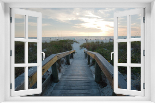 Fototapeta Naklejka Na Ścianę Okno 3D - Wood Crossover Bridge and Beach Access Staircase. View out to sunset near sunset . Shoreline, beach and blue sky with white clouds. Sea oats green on side with silhouette of people in background. 