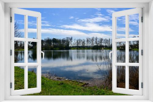 Fototapeta Naklejka Na Ścianę Okno 3D - Panorama photo of a large fish pond with reflection in the water, green grass and trees in the background against a blue sky.